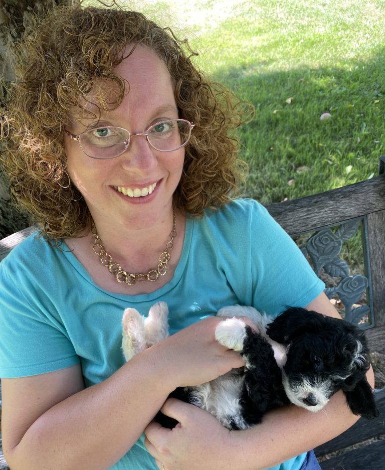 Amy of NY Dandy Doodles, woman holding black and white godlendooodle puppy sitting on bench under tree outside.