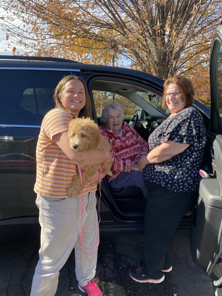 Happy family, 3 generations, grandmother, mother and daughter picking up their puppy from NY Dandy Doodles by their car with fall foliage around.