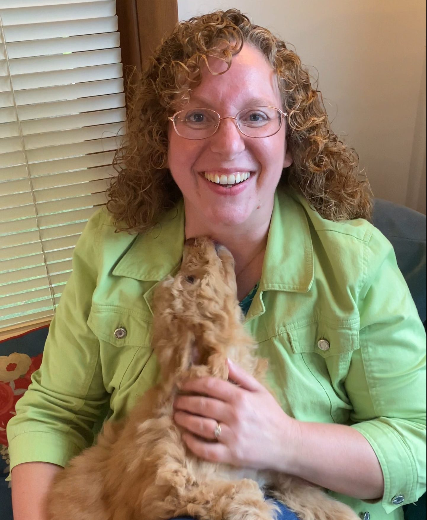 Amy, of NY Dandy Doodles, in a green jacket and holding a red fluffy goldendoodle puppy as it licks her neck.